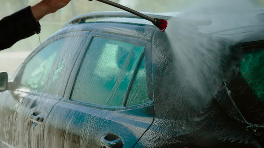 Unrecognizable woman rinsing side of car with high-pressure water at self-service wash. Female spraying vehicle windows and roof with strong water jet. Person cleaning automobile exterior using