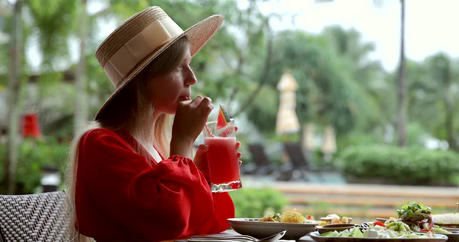 Young woman in summer hat enjoying tropical drink at outdoor cafe. Vacation dining, relaxation, travel concept.