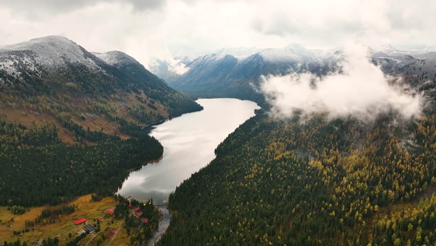 An aerial view captures a mountain resort with red-roofed buildings nestled by a serene lake, surrounded by dense coniferous forests and autumn foliage in a valley