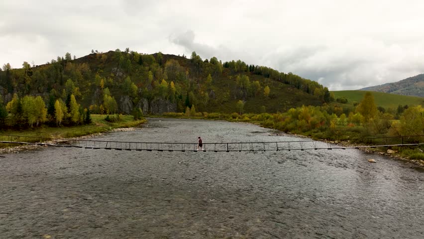 A lone person walks across a slender, low-hanging suspension bridge spanning a wide river, with a hillside of autumn-colored trees under a cloudy sky