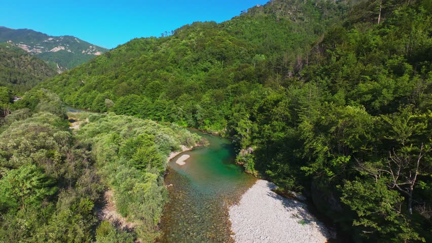 Aerial view of a crystal-clear green river flowing through a lush forest and mountain valley, with people relaxing on a pebble beach under a vibrant blue summer sky nature escape, calm relaxation
