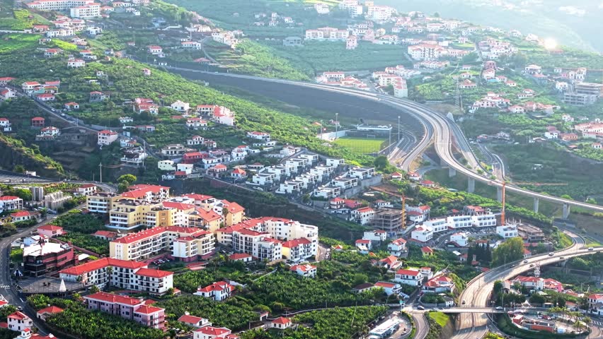 Aerial drone footage of the coastal city of Funchal, Madeira, Portugal. The video captures traditional red-roofed buildings, winding streets, and scenic mountain backdrops near the Atlantic Ocean.