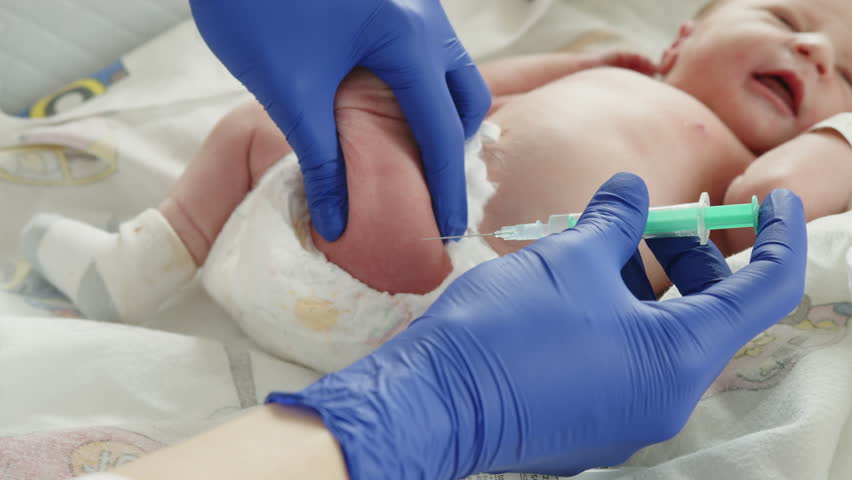 A newborn baby receives an injection from a medical professional in a hospital setting. The focus is on the vaccination process.