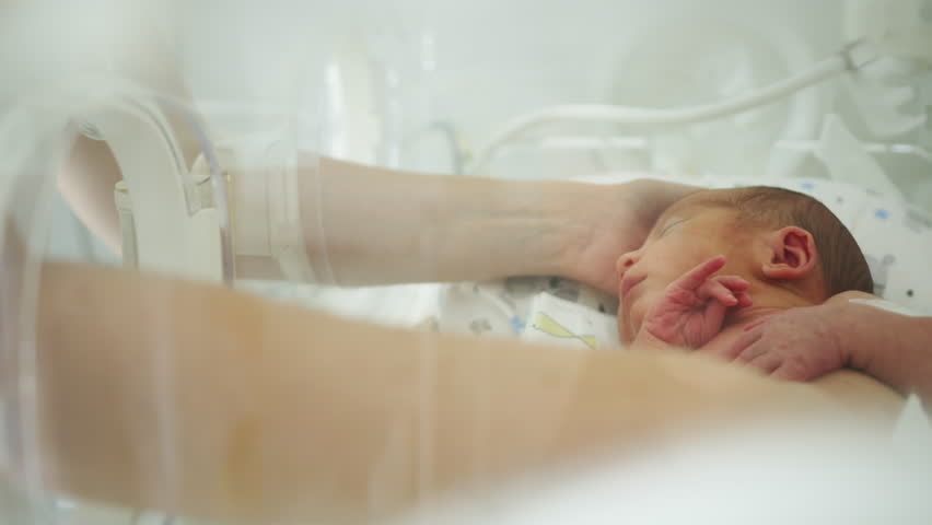 A premature baby in a neonatal incubator, with a caregiver's hands providing care. Medical equipment is also visible.