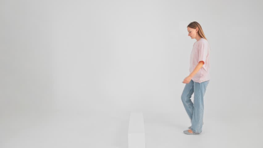 Woman is step on white curb and go ahead filmed in studio in slow motion. Isolated view of young adult Caucasian lady wearing jeans and pink T-shirt is overcoming stage. Theme of overpassing obstacle.