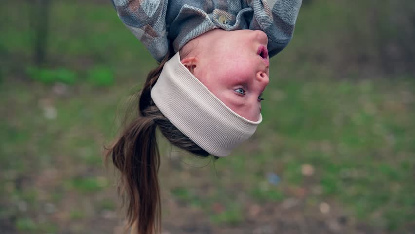Little schoolgirl is hanging upside down on a playground bar in the forest park and jumping filmed in slow motion. Closeup view of female Caucasian kid playing in woodland and doing exercises