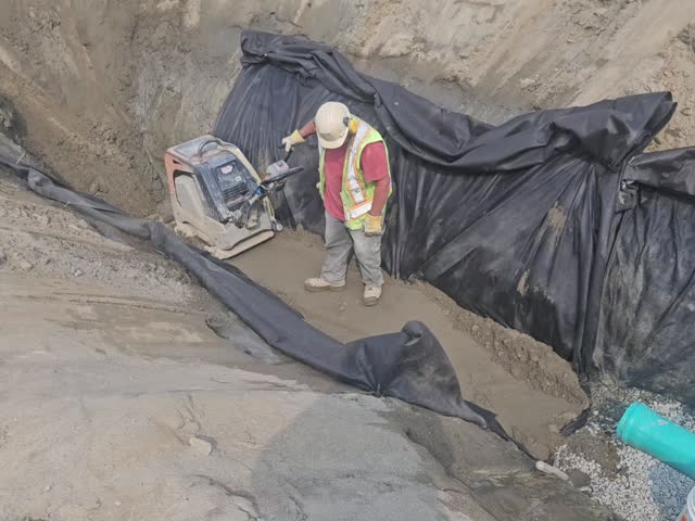 Laborer compacts trench subgrade using a reversible plate compactor over geotextile prior to placing bedding stone.
