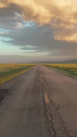 A long, cracked road stretches through a rural landscape in Carrizo Plain National Monument, California, USA, leading towards distant mountains under a dramatic, colorful sky at sunset.