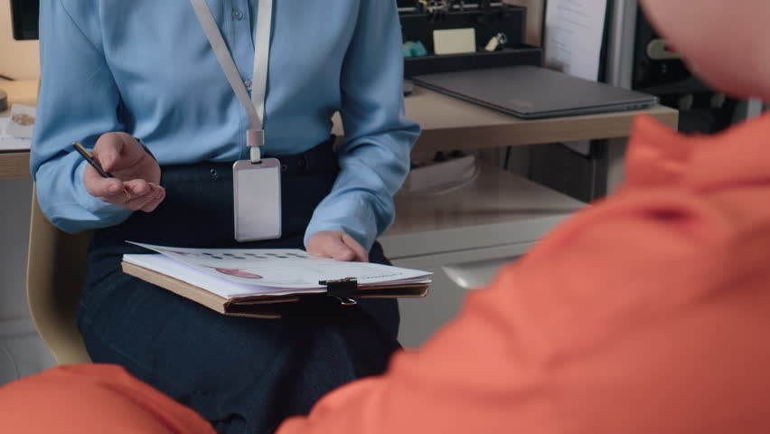 Zoom out handheld shot of female shaved head prisoner in orange jumpsuit communicating with middle-aged counselor analyzing criminal profile