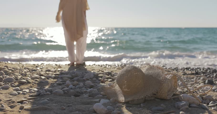 Stylish woman places down her straw bag and sun hat on a calm pebble beach, then walks toward the shimmering sea. Slow motion clip for wellness, travel, or natural lifestyle branding.