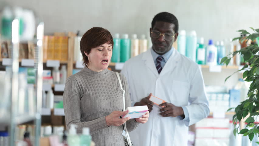 Woman choose buy painkiller tablet at pharmacy, compare with other good. Client check for absence of harmful substances, view product in details. African man pharmacist offer available assortment item