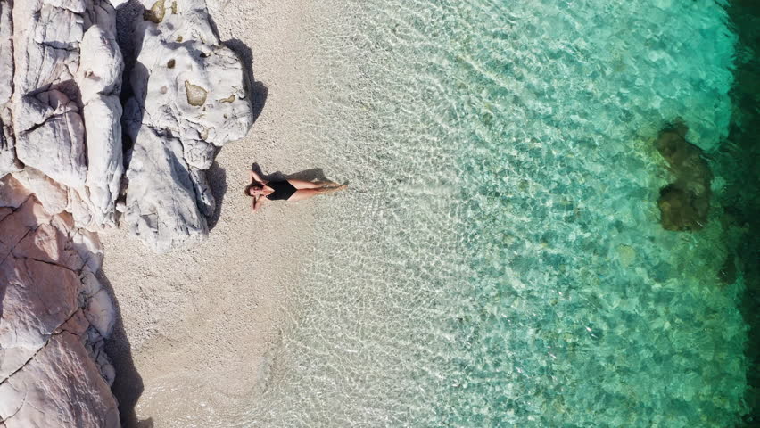 4K drone footage of woman dressed black swimsuit prostrated on white pebble beach lying down on the calm Ionian sea waves. Exotic tropical summer holidays concept. Cephalonia island, Greece.
