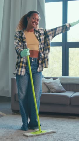 African American woman joyfully mops the floor in a spacious living room. She wears casual attire and dances while cleaning, radiating positive energy and happiness in a cozy space.