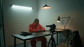 Medium long shot of African American male investigator in suit sitting down on chair next to video camera recording female Caucasian inmate in orange jumpsuit in interrogation room - Powered by Shutterstock - Get 15% off with code: PIKWIZARD15