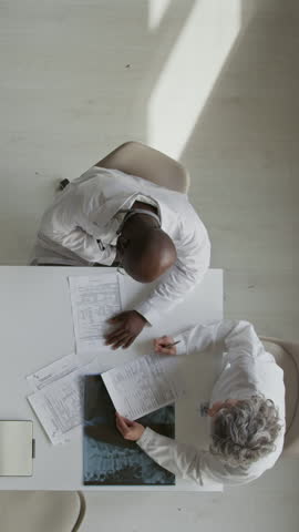 Vertical top view of two multiethnic male and female doctors sitting at table in office at medical clinic, reading patient history and treatment report, studying xray