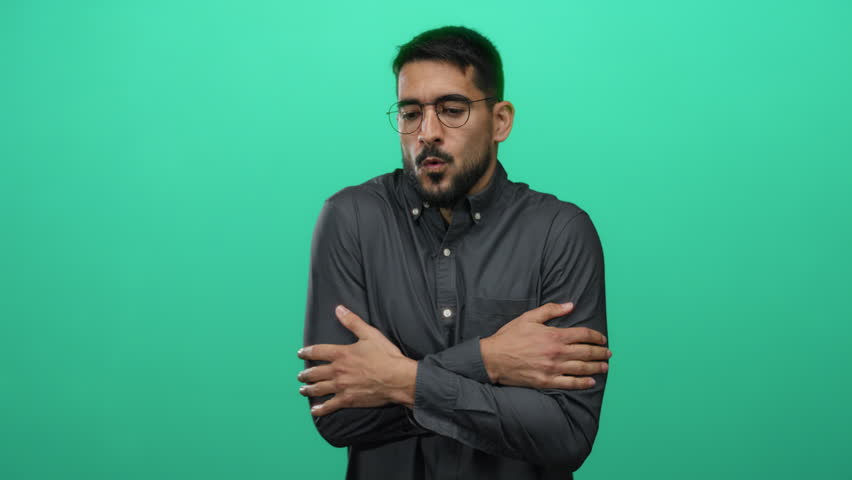 Young man shivering from cold in front of a vibrant green wall, portraying discomfort with a tense expression, arms crossed tightly, wearing glasses and a dark shirt. - Powered by Shutterstock - Get 15% off with code: PIKWIZARD15