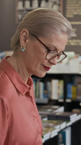 Vertical tilt shot of mature Caucasian woman arranging vibrant hardcovers on display table in cozy bookstore and taking her seat, preparing for personal autograph session and literary promotion