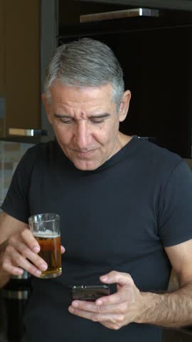 Middle-aged Man Relaxing at Home, Drinking Cold Beer and Scrolling on his Phone. The fit 54-year-old enjoys a free day at home, slowly drinks a chilled beer, and cheerfully browsing the phone.