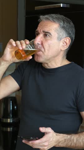 Middle-aged Man Relaxing at Home, Drinking Cold Beer and Scrolling on his Phone. The fit 54-year-old enjoys a free day at home, slowly drinks a chilled beer, and cheerfully browsing the phone.