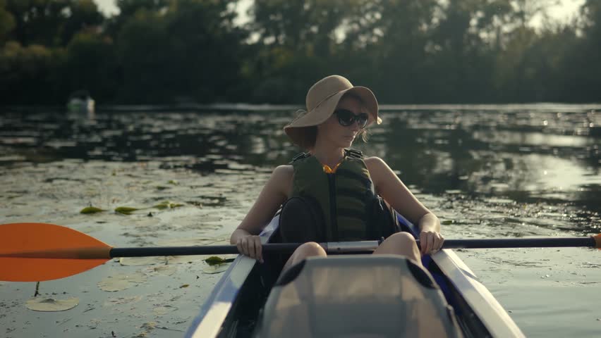 woman floating on kayak boat. beautiful woman in a sun hat and life vest paddles a kayak on a calm, scenic river. She is enjoying peaceful summer vacation and an active outdoor lifestyle on sunny day