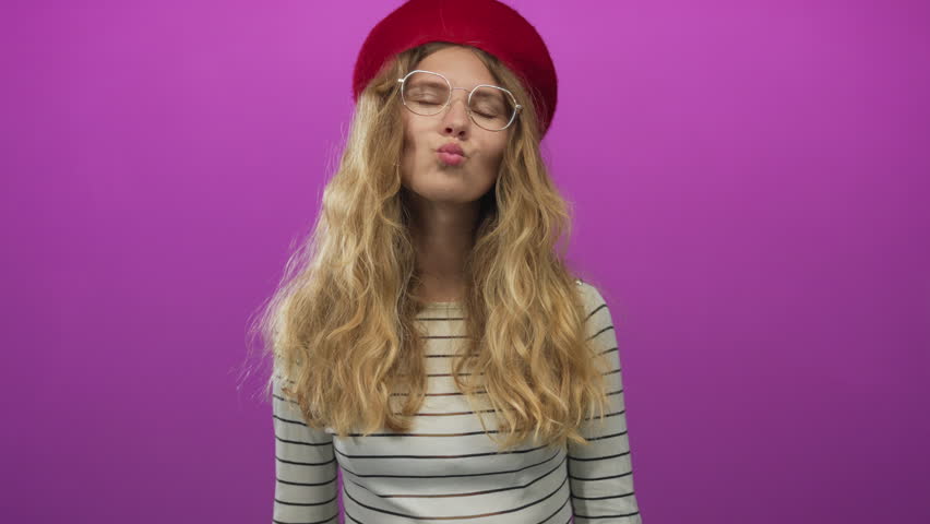 Woman uses fingers to form a heart gesture in a studio scene with blonde young model smiling against a purple backdrop wearing fashion casual attire.