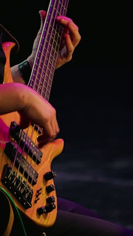 Slow motion close-up of a man playing bass guitar on a dark stage. Bass guitar has five strings and stylish wooden body