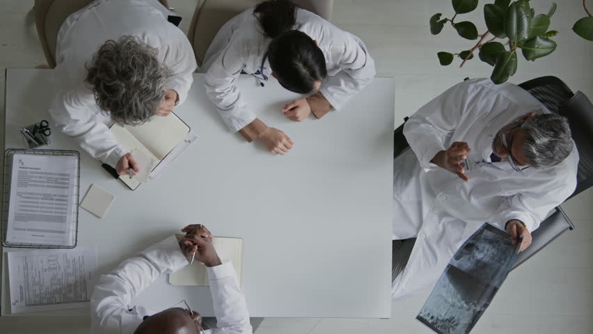 Top shot of four multiethnic male and female doctors in white coats sitting around table in hospital office, frustrated surgeon demonstrating xray of patients spine to colleagues, asking for opinion