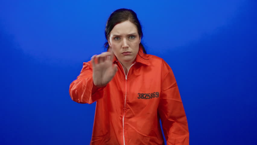 Woman in orange prison jumpsuit against blue background expresses frustration covering ears and gesturing, highlighting emotional intensity in a confined setting.