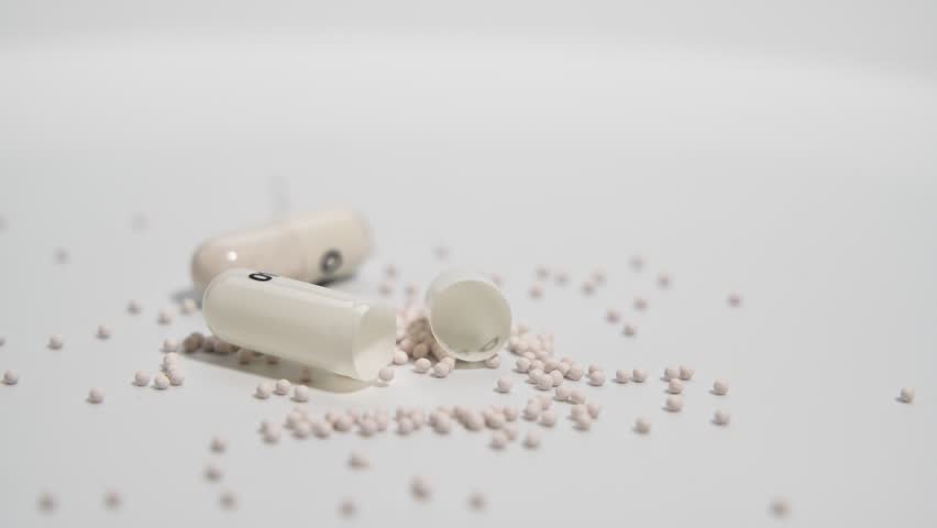 Open Capsules with Granules on White Surface. Capsules lie open on a smooth surface, showing fine granules spilling out while the camera rotates slowly. Detailed pharmaceutical composition is the focu