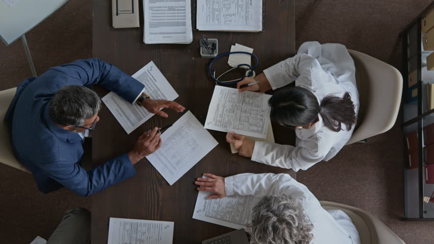 Top zoom-in shot of male medical clinic director in blue business suit talking to staff doctors during daily meeting, studying clinical recommendations on various drugs for patient treatment