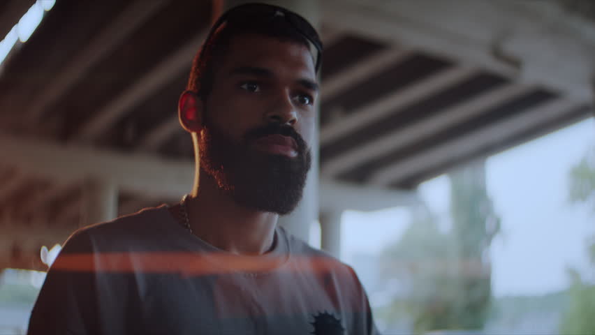 Tranquil African American man with stylish beard stands under concrete overpass bridge at backlit. Young black male with thoughtful expression in city setting