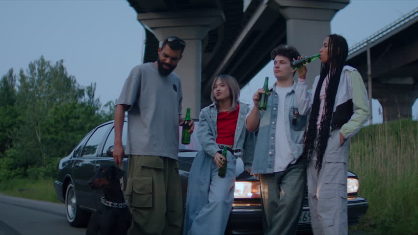 Group of young people stand near car under overpass with bottles of beer engaged in social gathering. Urban environment transitions into natural surroundings