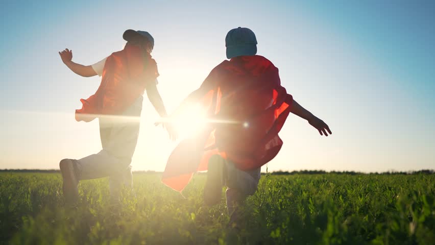 Two people in field with red cape. Children running in the park playing superheroes. Young boy with fun imagination in a field. A pair of individuals in a meadow with a crimson cape lifestyle.