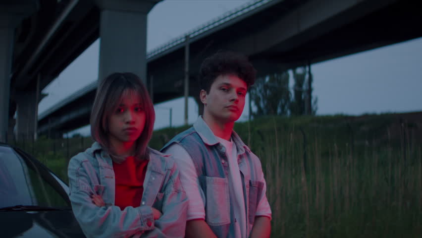 Woman and man stand with confident expressions near car under structures of overpass. Contemplative gaze and urban setting evoke edgy aspect of youth culture