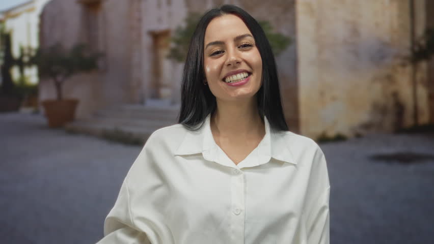 Young hispanic woman smiling on an old town street holding a credit card, showcasing happiness and elegance in a charming outdoor setting with historical architecture.