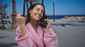 Woman in pink robe holding vintage phone and applying makeup by the seaside promenade, showcasing a joyful moment against a sunny beach backdrop with palm trees and ocean view. - Powered by Shutterstock - Get 15% off with code: PIKWIZARD15