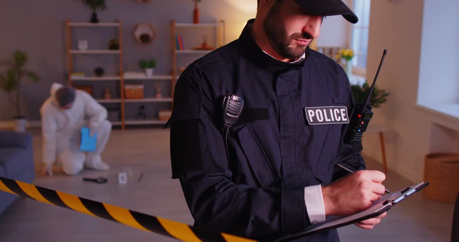 Cropped view of male police officer standing near caution tape, gathering evidences at crime scene. Policeman writing down details for report with forensic scientist investigating crime on background.