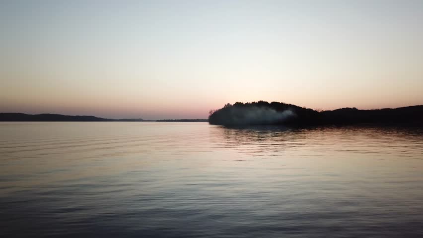 Smoke drifts from a wooded peninsula into the calm waters of Lake Wisconsin under a soft twilight sky. The wide aerial view highlights serene reflections and a mysterious dusk atmosphere.