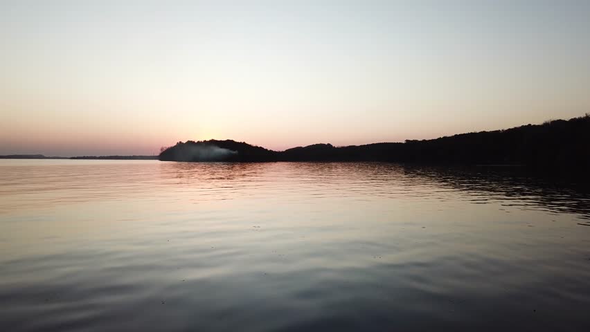 As dusk settles over Lake Wisconsin, a soft gradient sky reflects on the smooth water. Smoke drifts from a wooded peninsula, creating a tranquil and moody lakeside atmosphere.