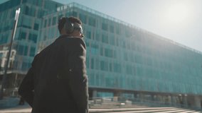 Stylish black man walking down city street and listening to music by headphones. Self-confident passerby in black coat and sunglasses admiring modern architecture, enjoying city life in metropolis - Powered by Shutterstock - Get 15% off with code: PIKWIZARD15