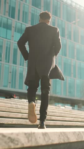 Cinematic shot in urbanscape, man running up of upstairs in business district. Back view of stylish african male person in black coat moving to office building, move to future success, career growth