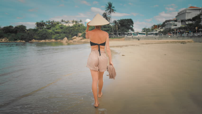 Graceful woman in bikini, shorts and conical hat walking on seashore in Vietnam. Back view of lady strolling alone on beach, happy and carefree female vacationer enjoying simple life, traveling