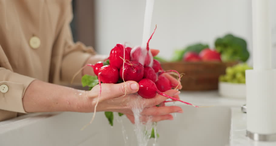 Slow-motion footage of woman's hands are washed a bunch of red radishes under the pressure of flowing water in the white kitchen, a basket with tomato, broccoli and onions in the background, grapes