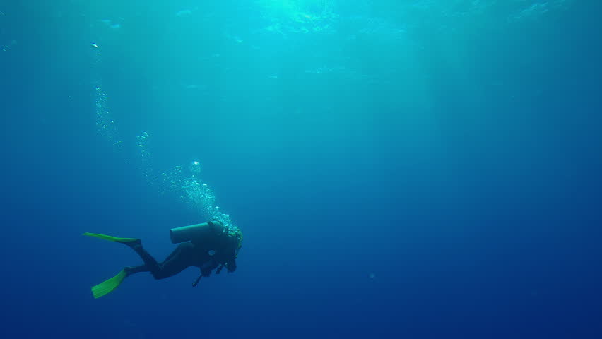 Scuba diver swims in the blue depths of the ocean illuminated by sunrays, Back light, Slow motion of a diver swims on the deep in sunlight, contre-jour