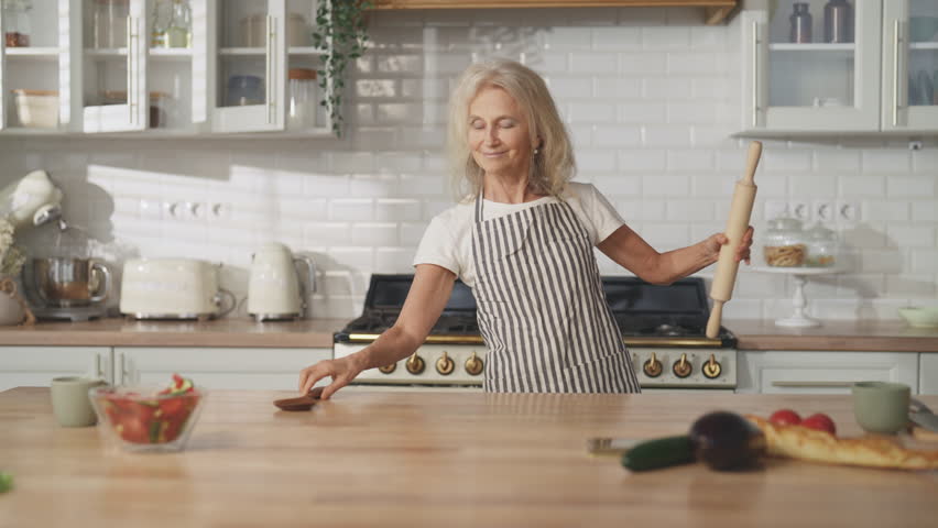 Happy old lady cooking at home and dancing, medium portrait in kitchen, have fun . Cute mature woman dancing and listening to music when doing housework, recharging energy, good mood and positivity