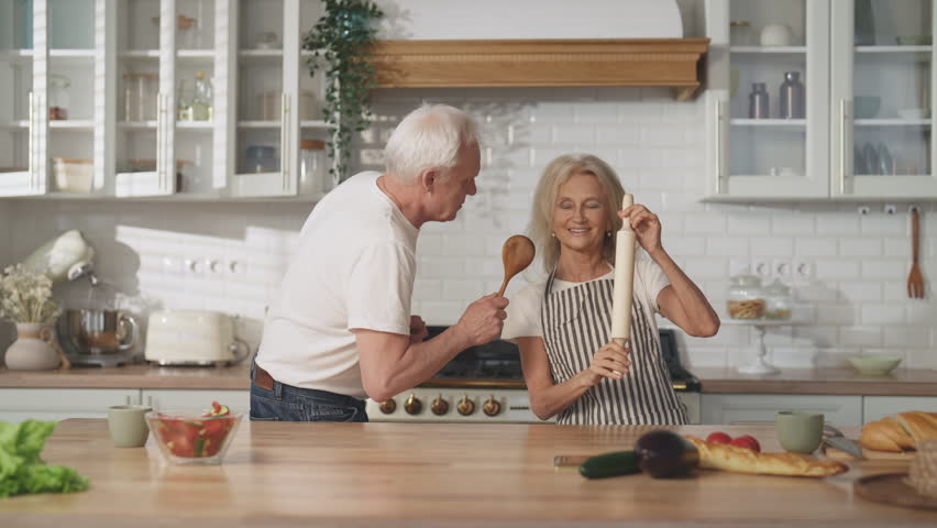 Enjoy simple life and be happy, old wife and husband dancing in home kitchen. Elderly lady singing into rolling-pin, aged man dancing with wooden spoon, fun and joy, merry weekend of senior spouses