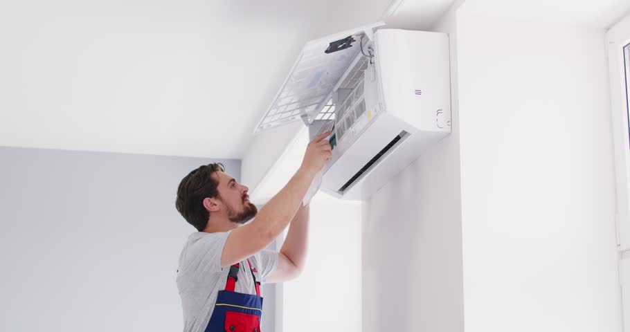 Worker from AC service repairs a modern air conditioner on the wall at home. Young man in a uniform uses a screwdriver while fixing the inside parts of an air conditioning system