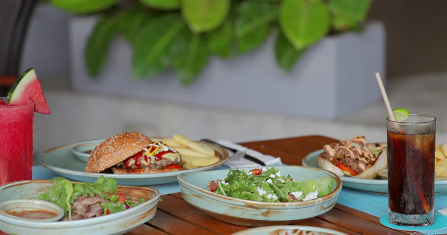 Casual outdoor dining scene with burger, salad, fries, and drinks on wooden table. Green plant background. Perfect for summer dining, food lovers, restaurant themes.