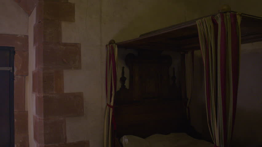 Overview of a dimly lit medieval-style bedroom with a carved wooden canopy bed, draped with red and green striped curtains, and a wooden cradle, set against thick stone walls and a shuttered window.
