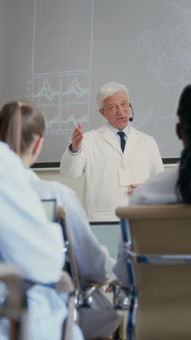 Vertical shot of male clinical professor in lab coat equipped with earset microphone delivering lecture to colleagues on latest developments in medical technology during conference in auditorium